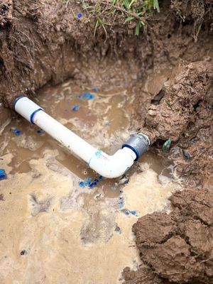 Leaking white PVC pipe elbow in a muddy dirt trench with standing water