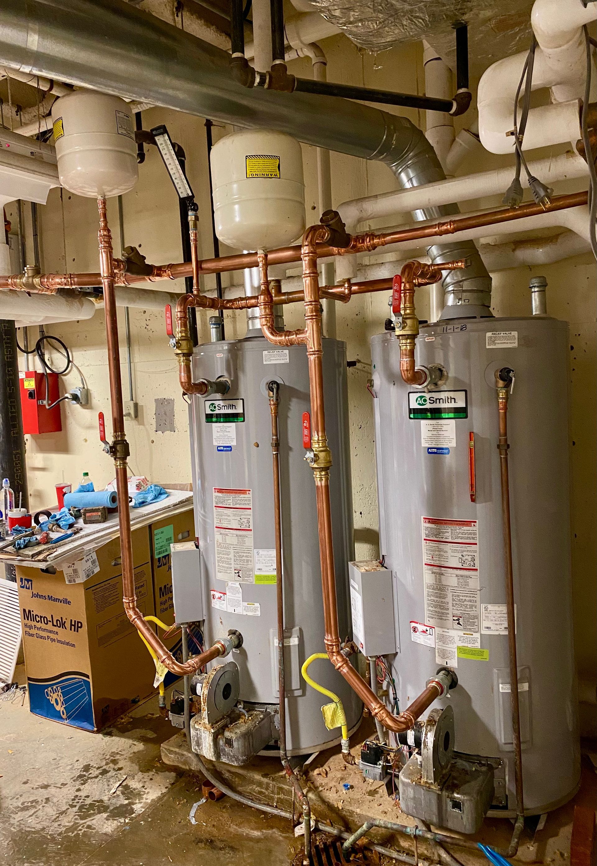 Two gray water heaters in a utility room with exposed pipes and ductwork
