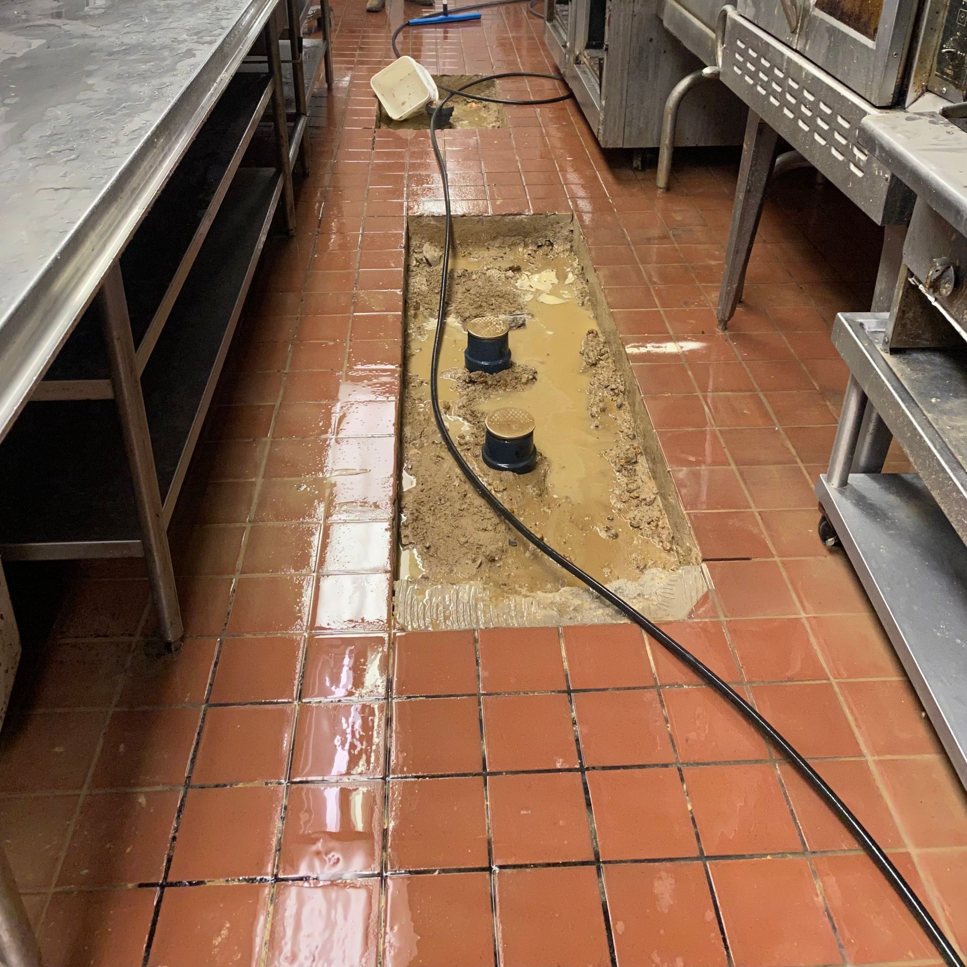 Commercial kitchen floor with a wet, damaged tile trench and a black hose lying across it