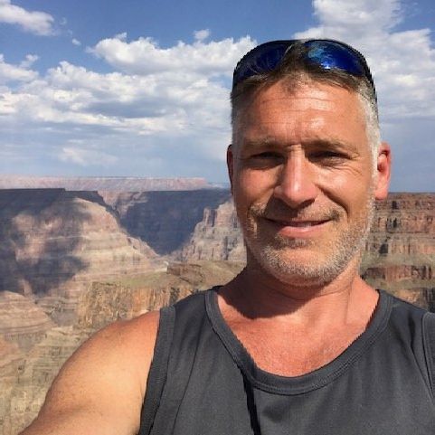 A man wearing sunglasses is taking a selfie in front of a canyon.