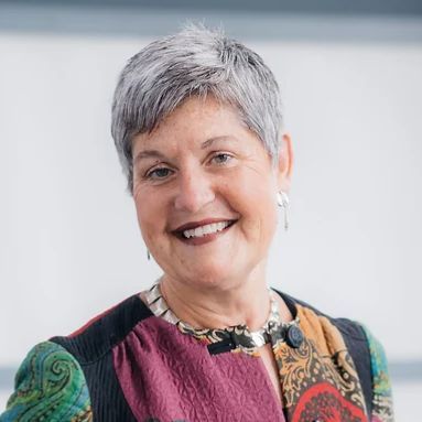A woman with gray hair is smiling for the camera while wearing a colorful jacket and earrings.