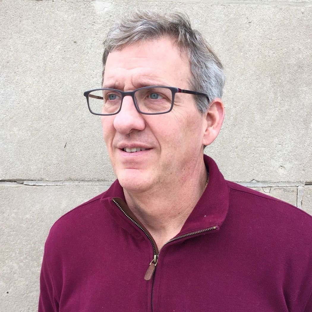 A man wearing glasses and a maroon sweater is standing in front of a brick wall.
