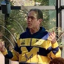 A man wearing a blue and yellow striped shirt with the letter m on it is standing in front of a window.