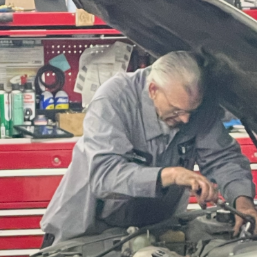 Mechanic working on a car engine, wearing a gray uniform, in a garage with red cabinets and tools.