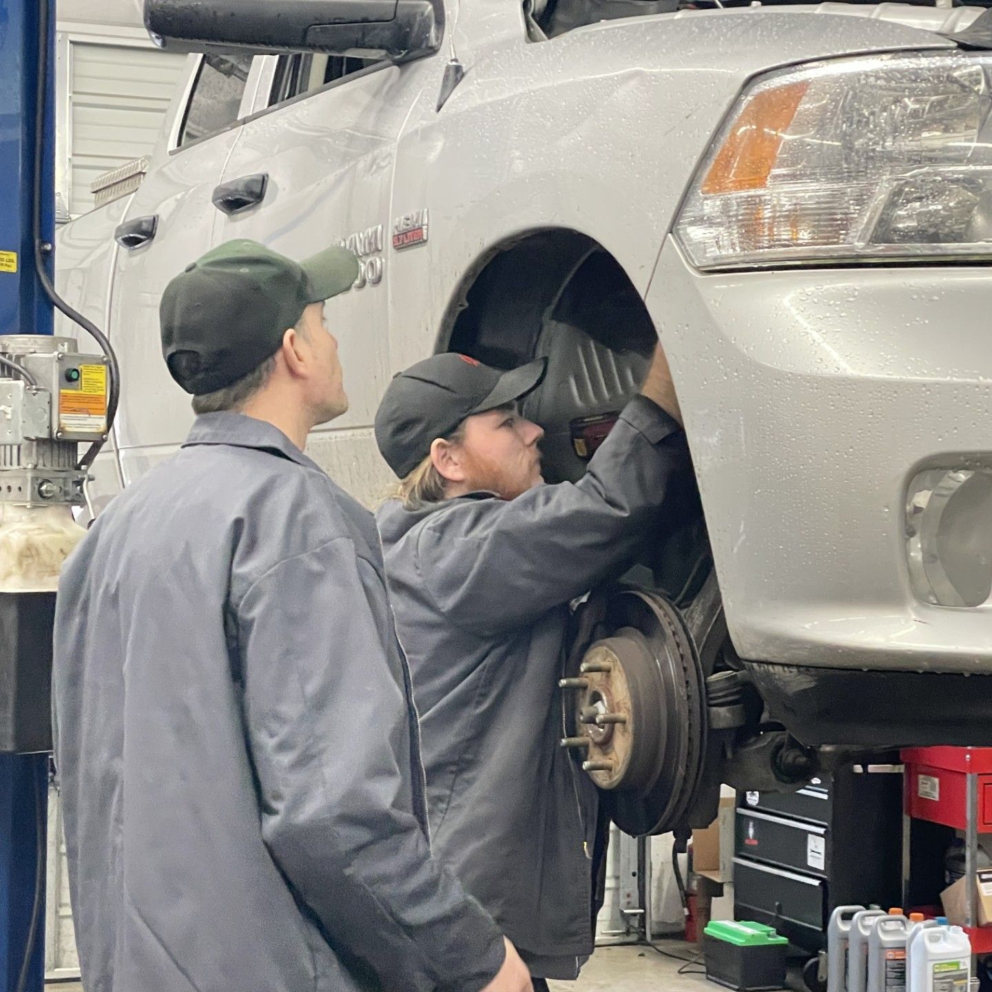 Two auto mechanics working on a silver truck in a repair shop, one looking on, the other working on the wheel.