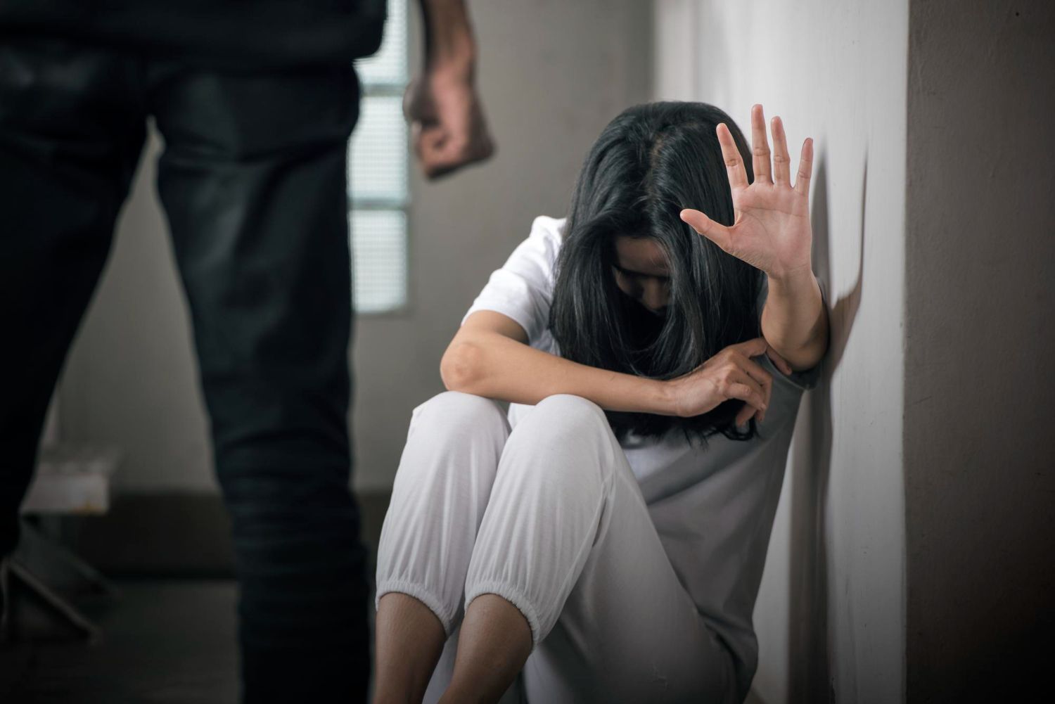 Person sitting on the floor, shielding face with raised hand as another person stands nearby in a dim hallway.