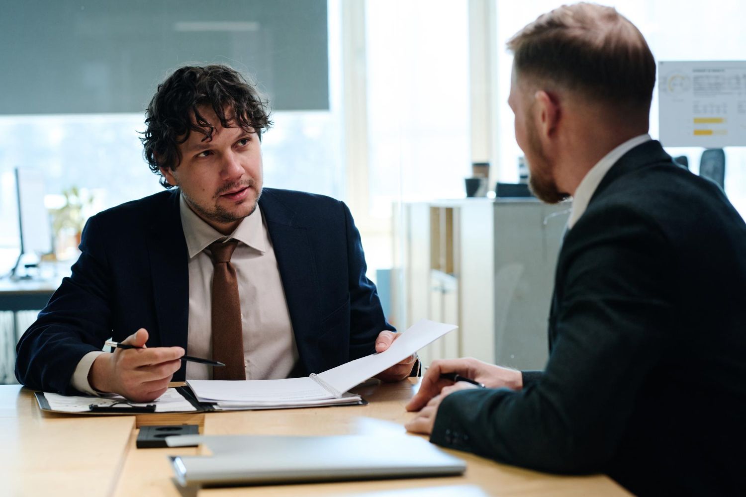 Two professionals in a business meeting, discussing documents at a desk in a bright office.