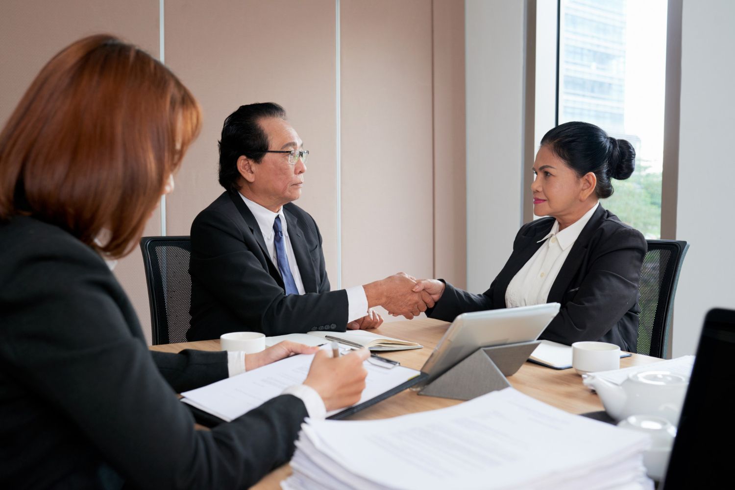 Three people in business attire shake hands across a conference table with papers and a laptop.