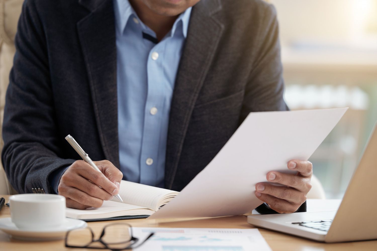Person in a blazer writing on paper and holding a document at a desk with coffee and glasses.