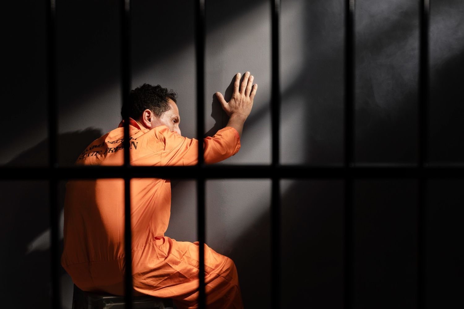 Person in orange prison jumpsuit kneels behind bars, hands raised against a dark wall.