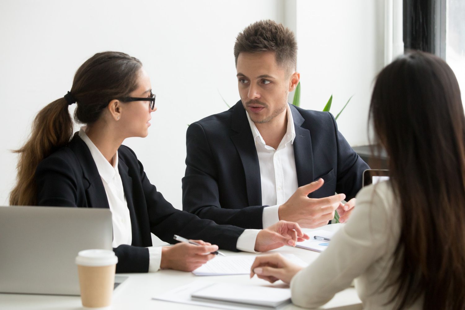 Three people in business attire discuss documents at a table in a bright office.