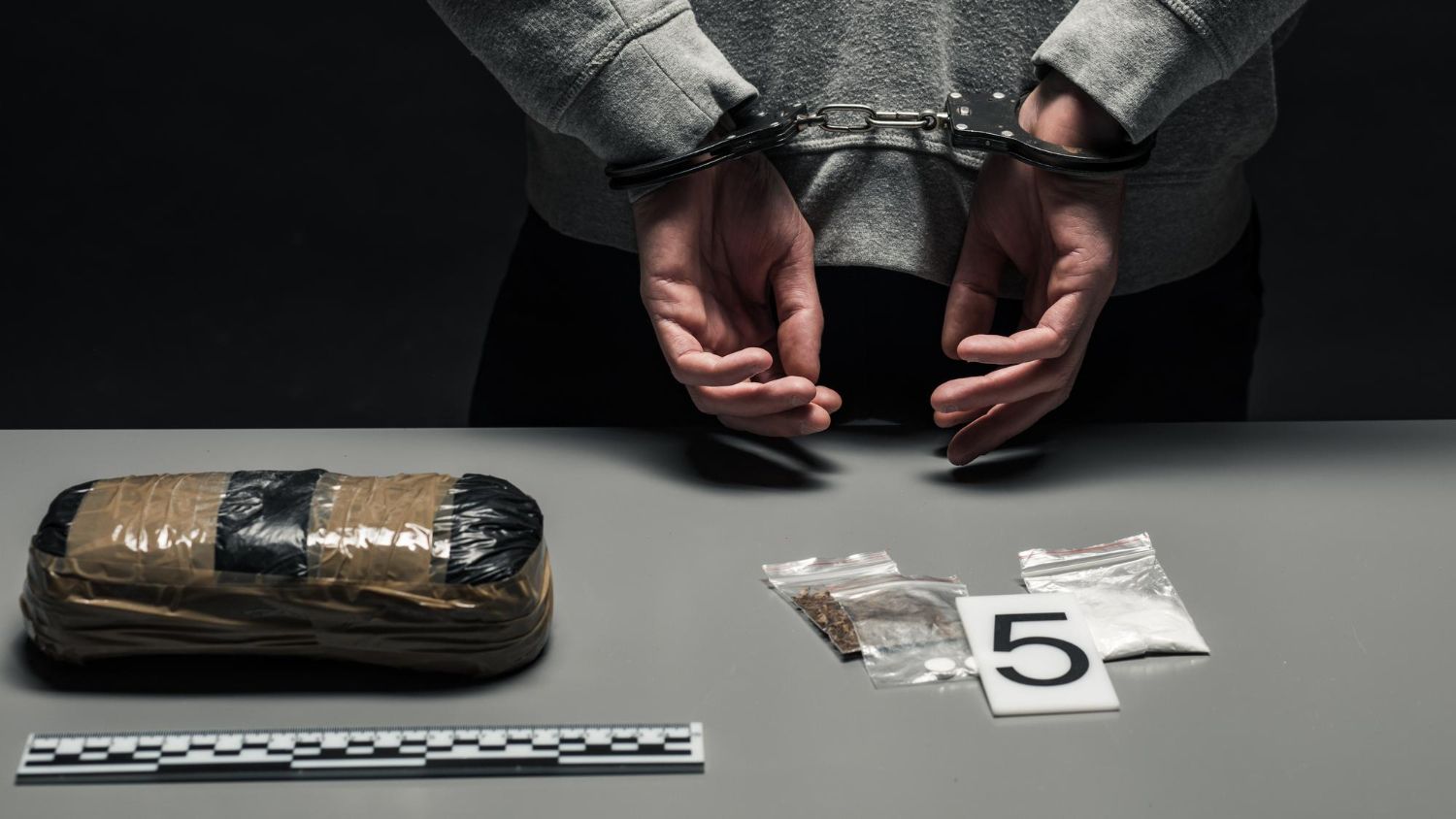 Handcuffed person behind table with wrapped package, scale ruler, and evidence marker 5