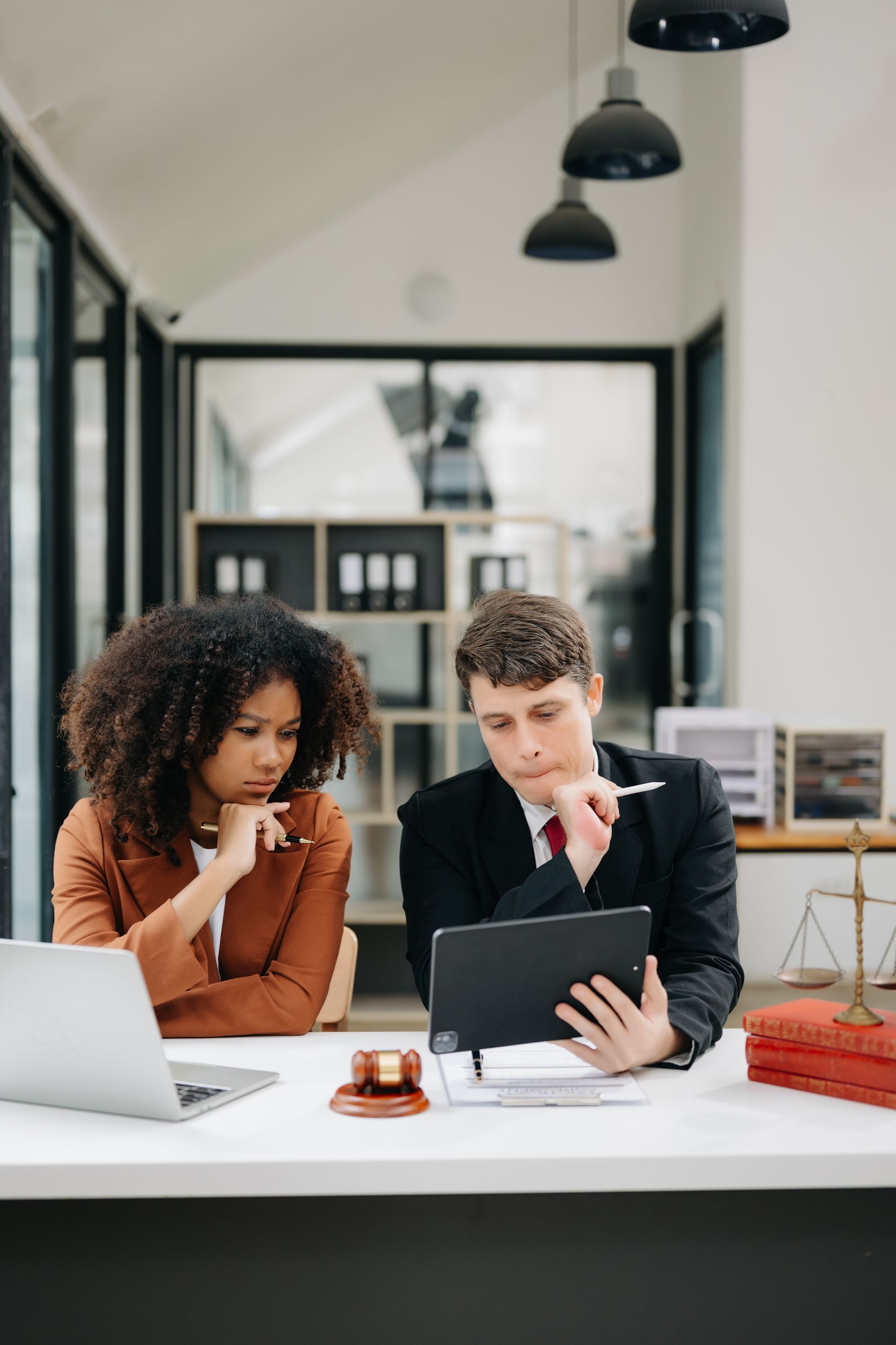 Two coworkers reviewing documents at a desk in a bright office, with a laptop, tablet, and coffee cup.