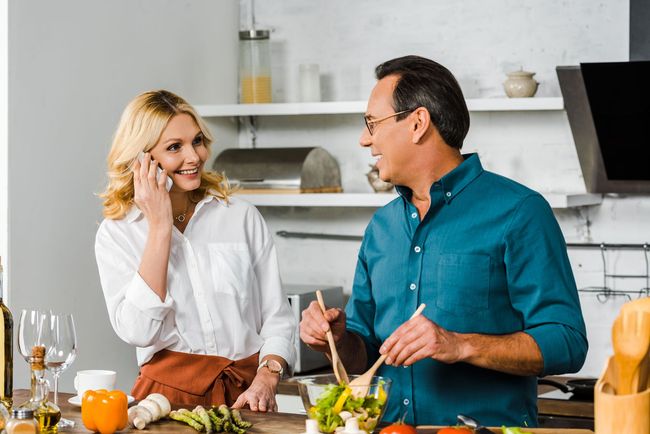 Two people cooking together in a bright kitchen, smiling and chatting over fresh ingredients.