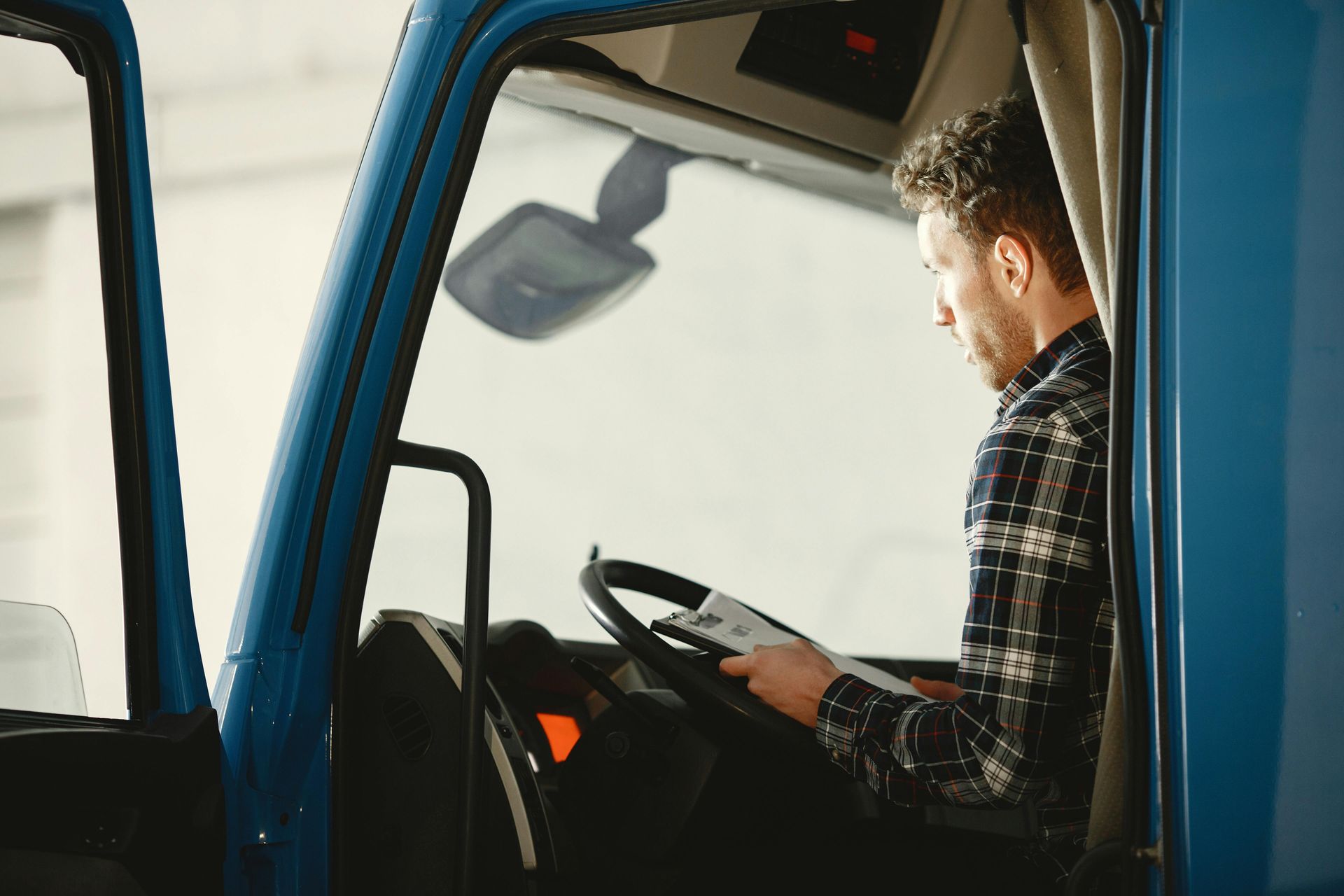 Man seated in a blue vehicle cab, looking at the steering wheel while holding a tablet