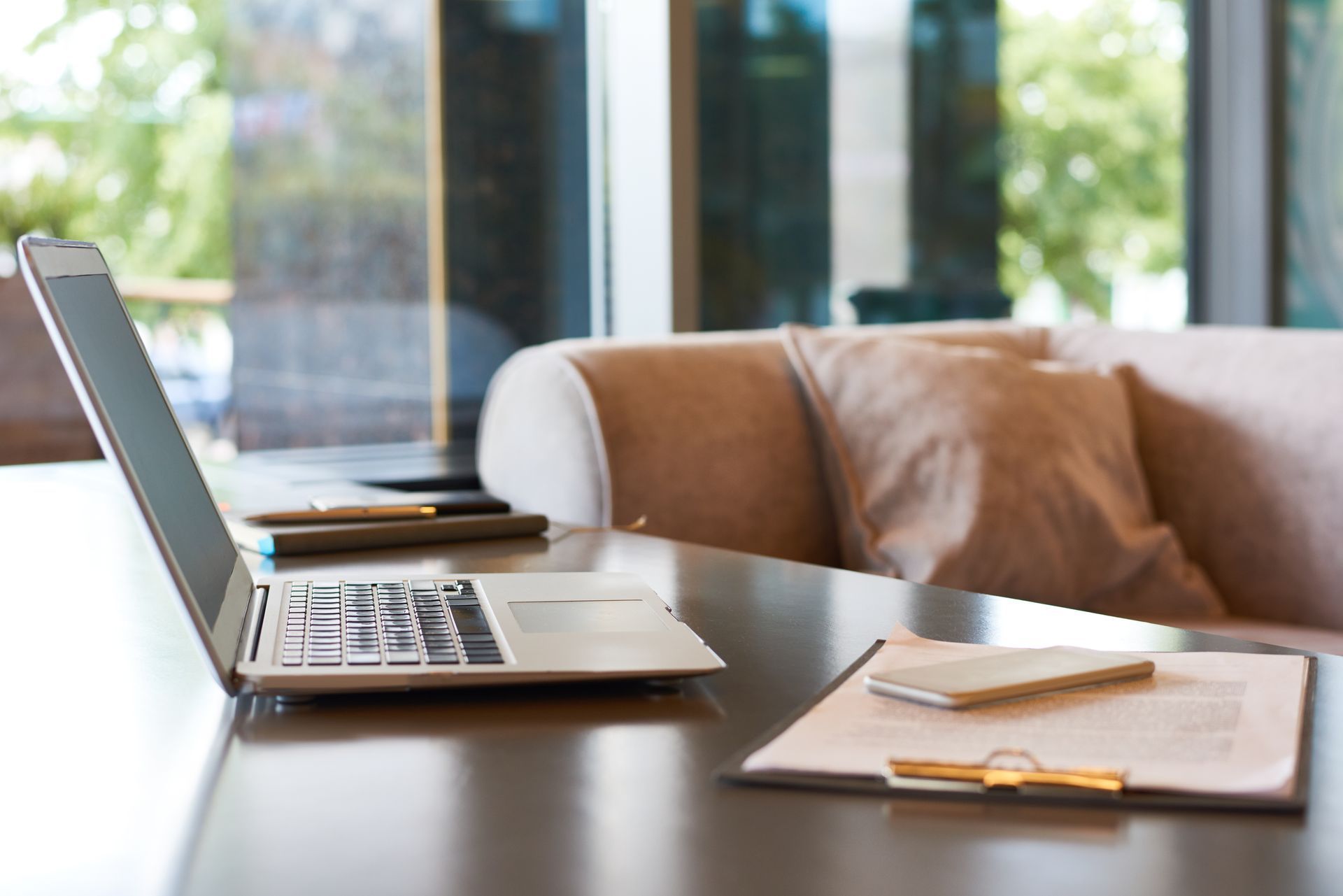 Laptop and notebook on a wooden table beside a couch in a bright office lounge