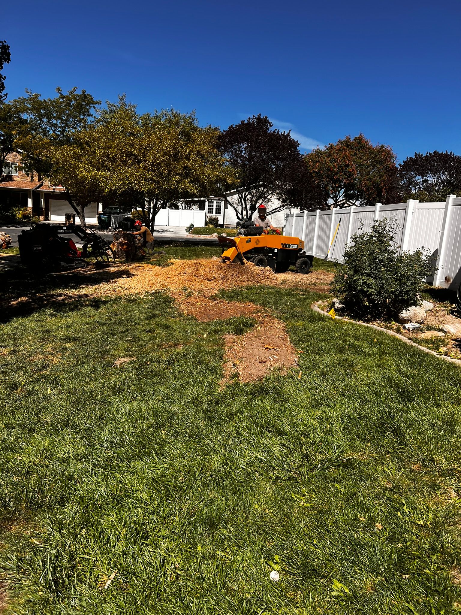 A stump grinder in a yard with a freshly ground stump, surrounded by grass, trees, and a white fence.