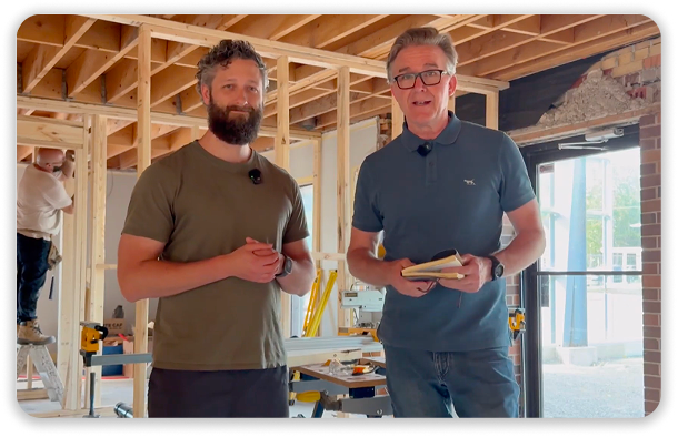 Two men holding phones stand in a partially built wooden interior with exposed framing and large windows.