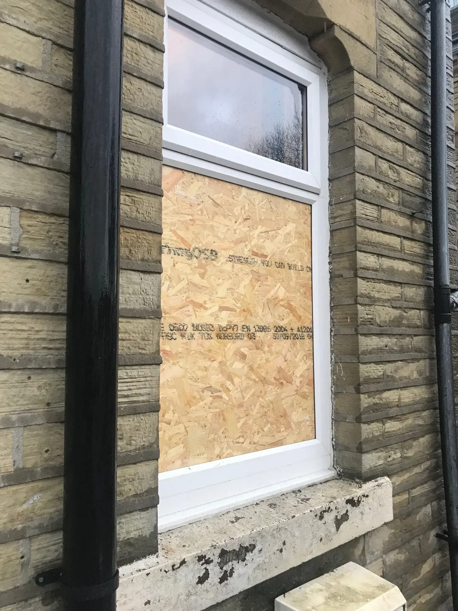 Window with boarded-up section; weathered brick wall; white frame; black downspout; exterior.