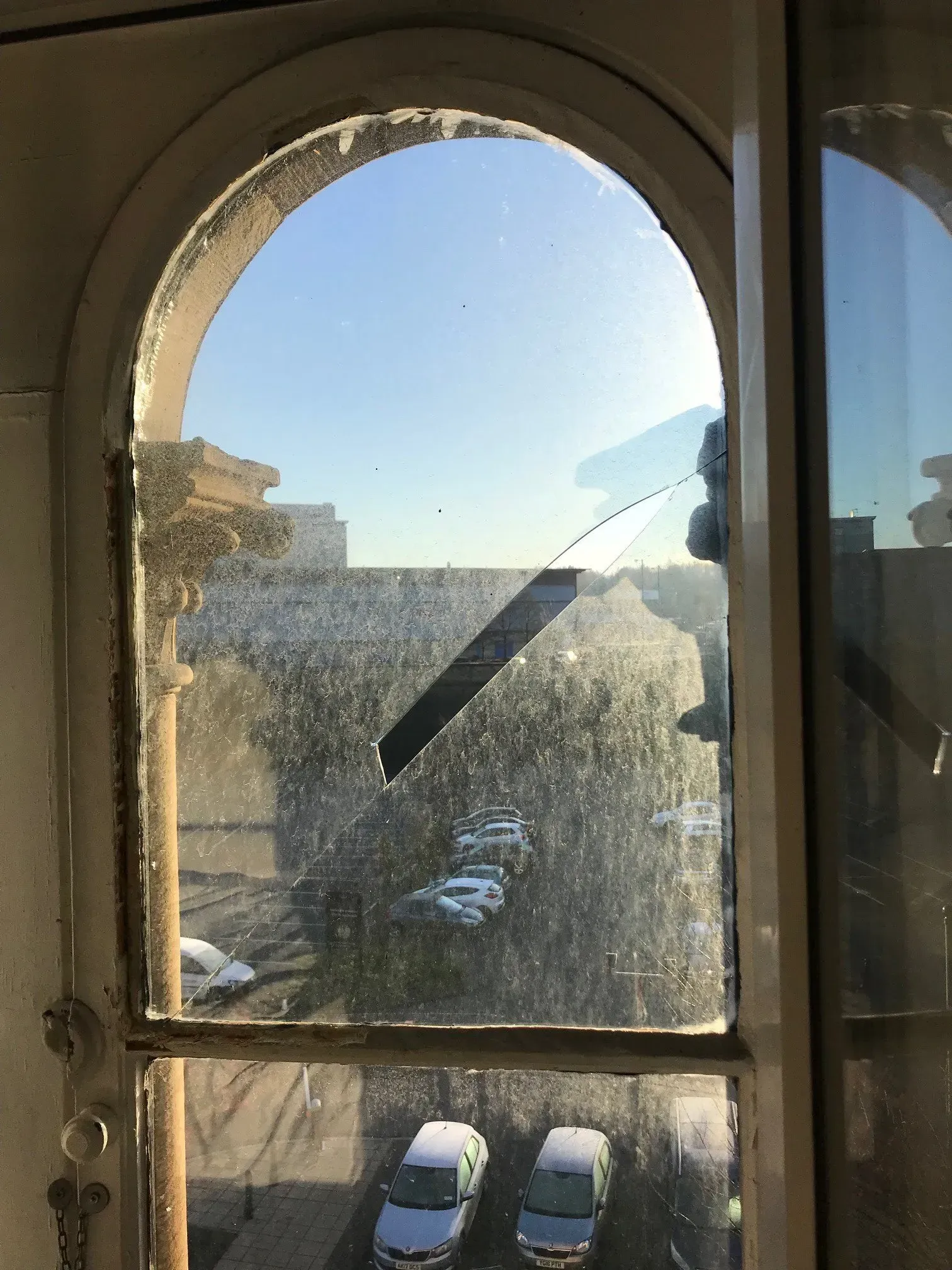 Arched window with cracked glass, overlooking a street with parked cars on a sunny day.