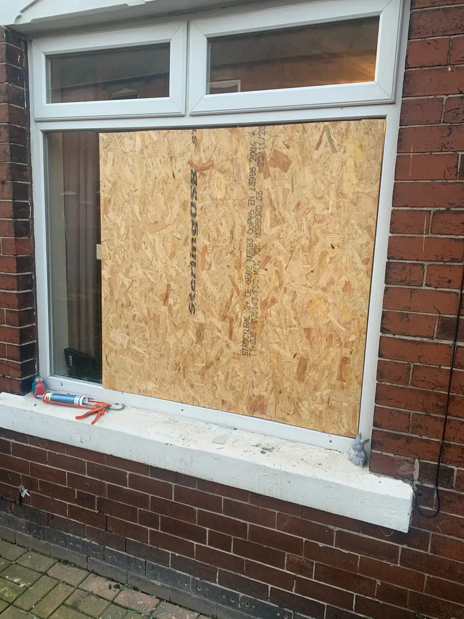 Exterior view of a window with the glass pane replaced by a wooden board; brick wall.