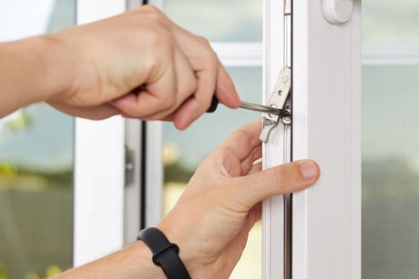 Hands using screwdriver to adjust window latch on white window frame.