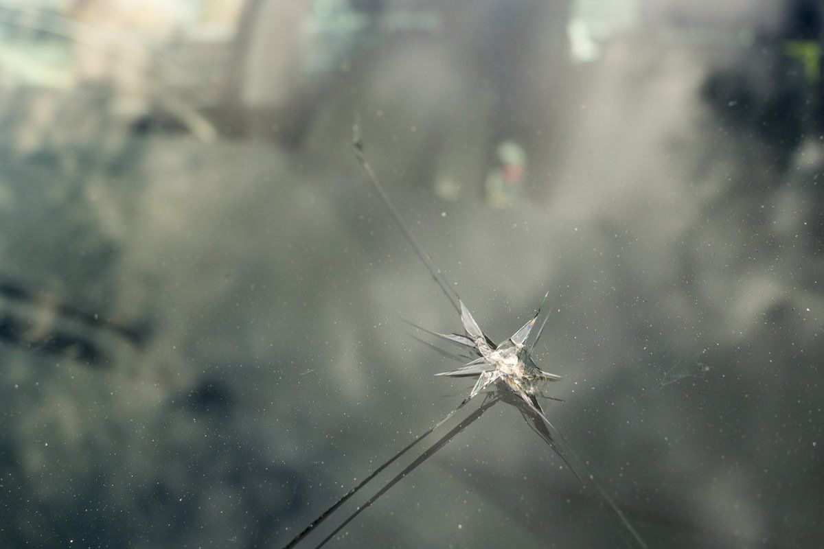Close-up of a cracked windshield. Crack emanates from a central point, with visible streaks. Background blurred.