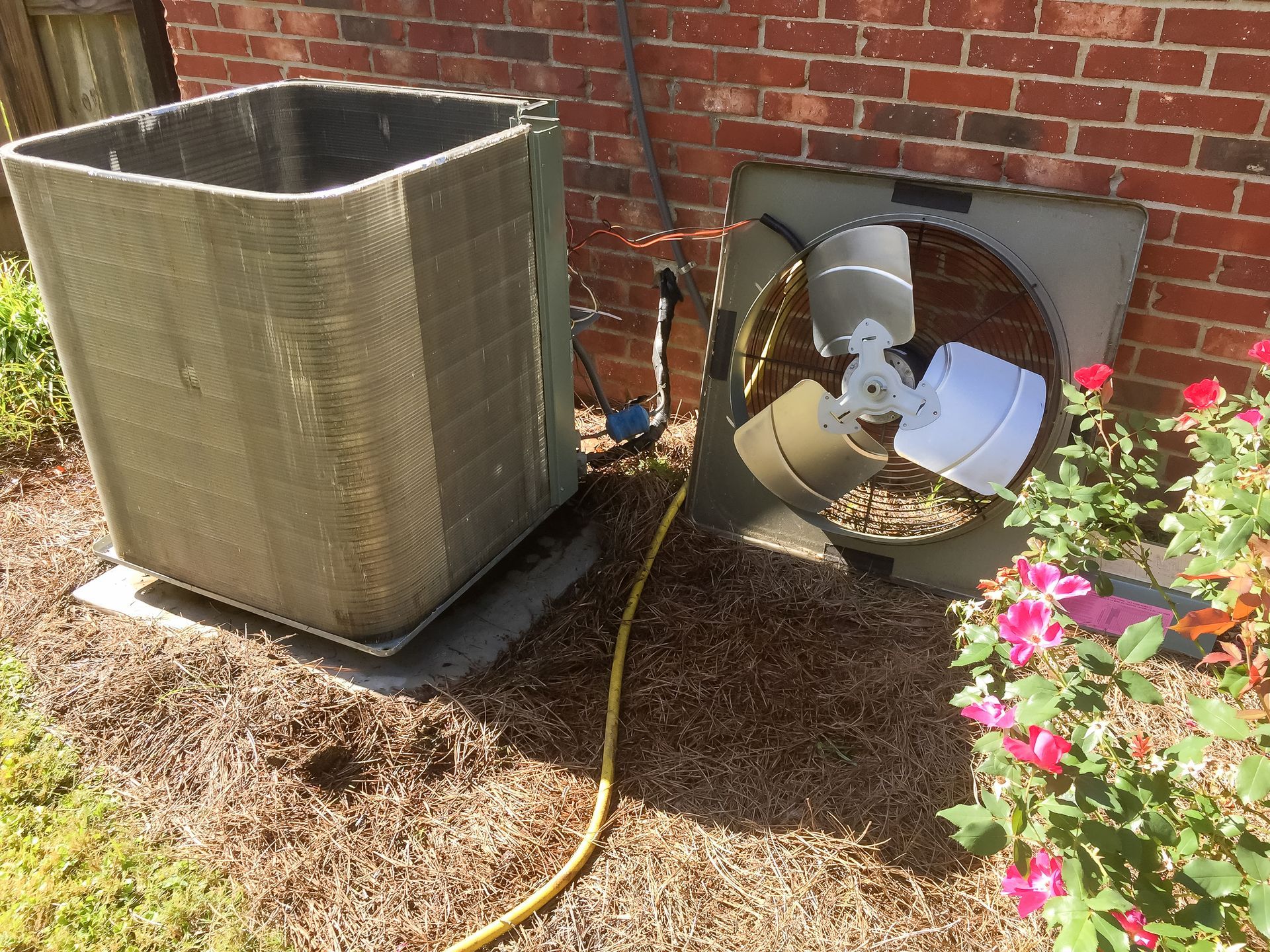 AC unit with fan disassembled next to a brick wall and plants in an outdoor setting.
