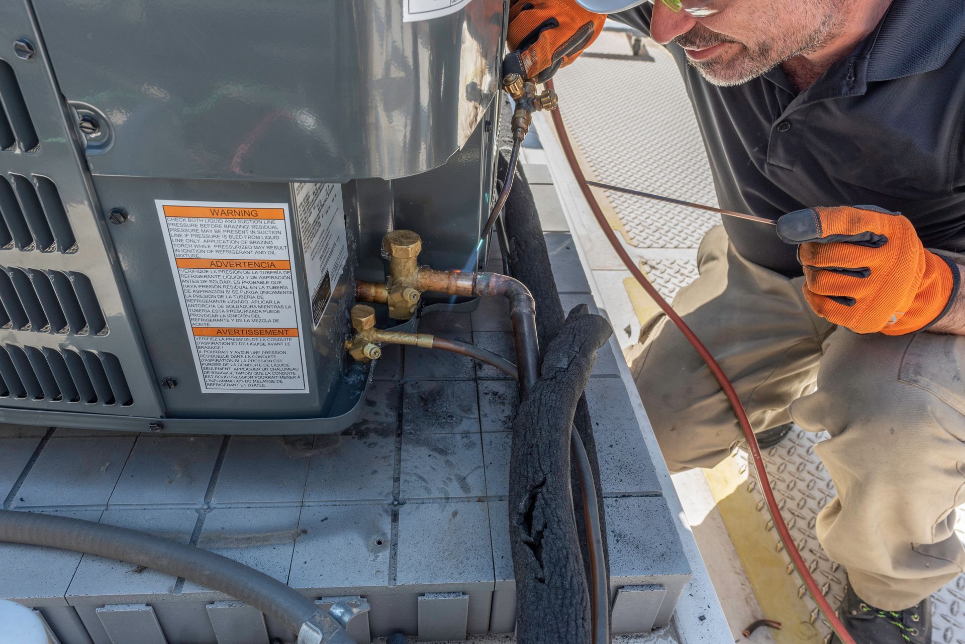 HVAC technician working on an air conditioning unit outside. He wears orange gloves and safety glasses.