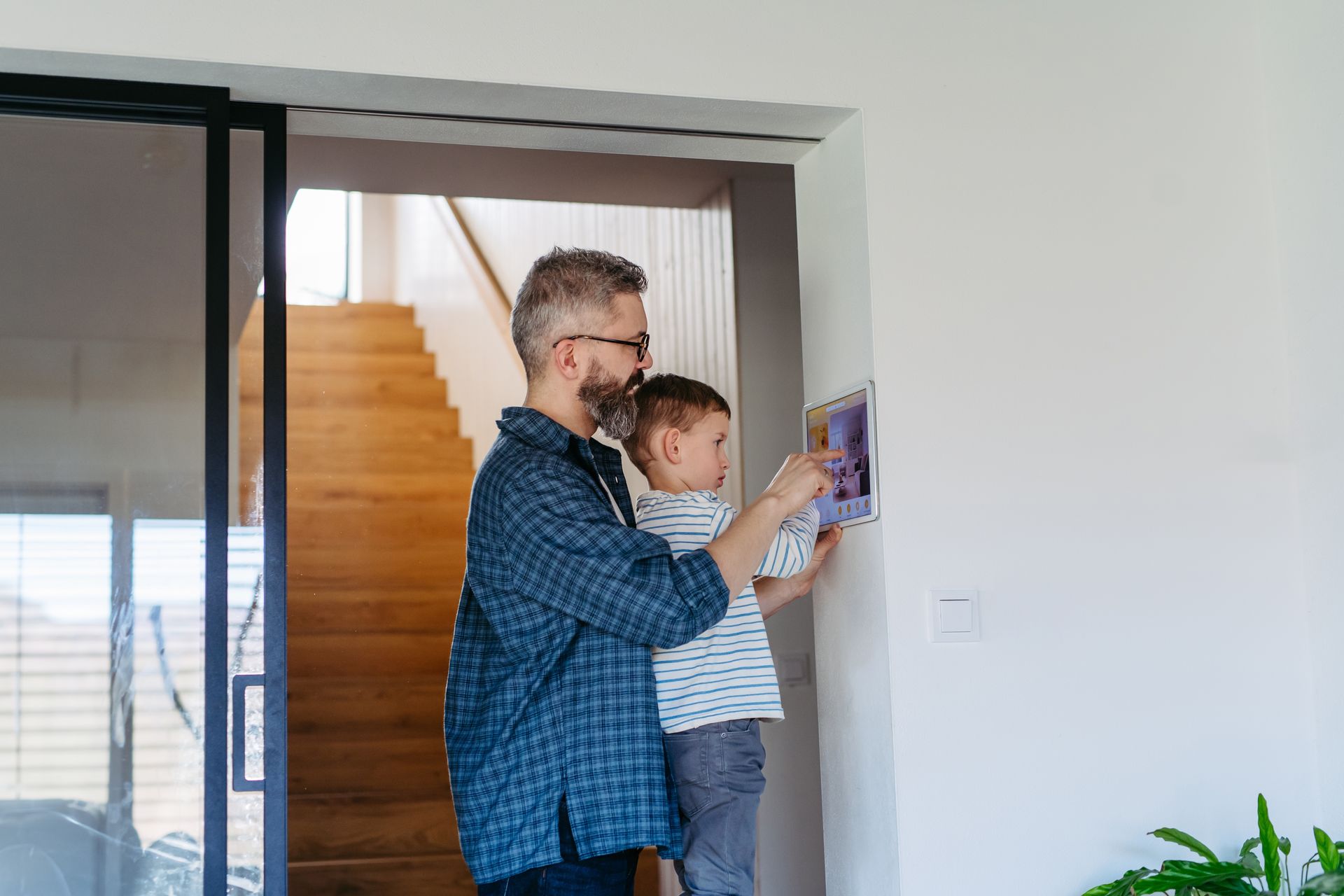 Man and child interact with a wall-mounted tablet near a staircase and doorway.