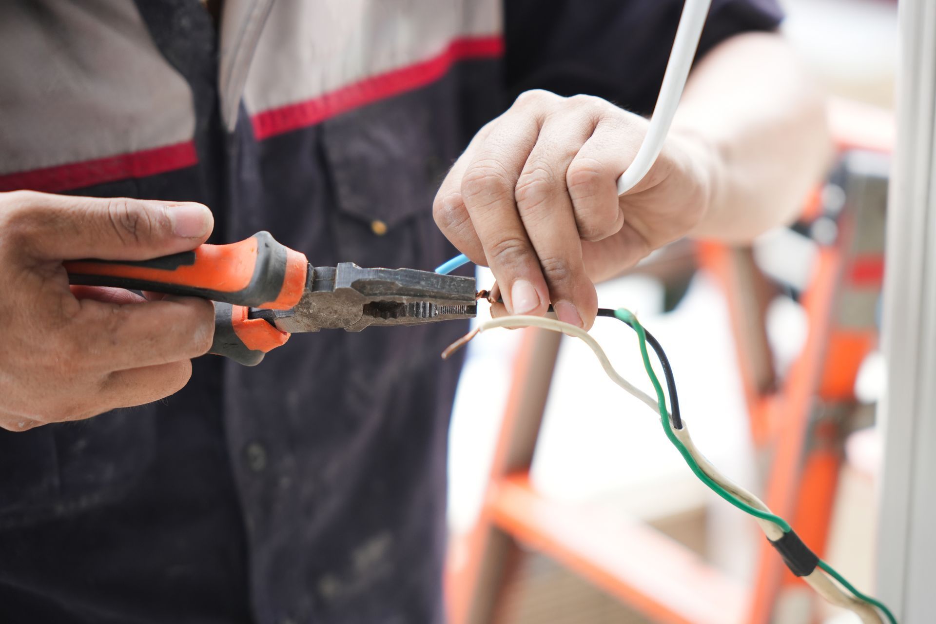 Electrician using pliers to strip wires near an orange ladder.