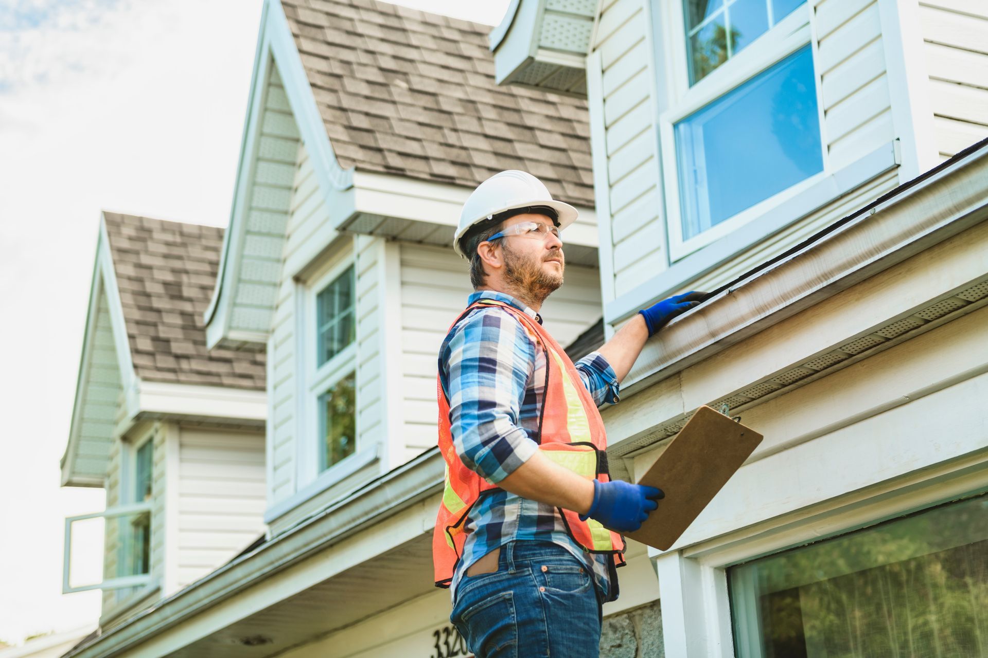 A construction worker in safety gear inspects a house's siding, holding a clipboard and looking upwards.