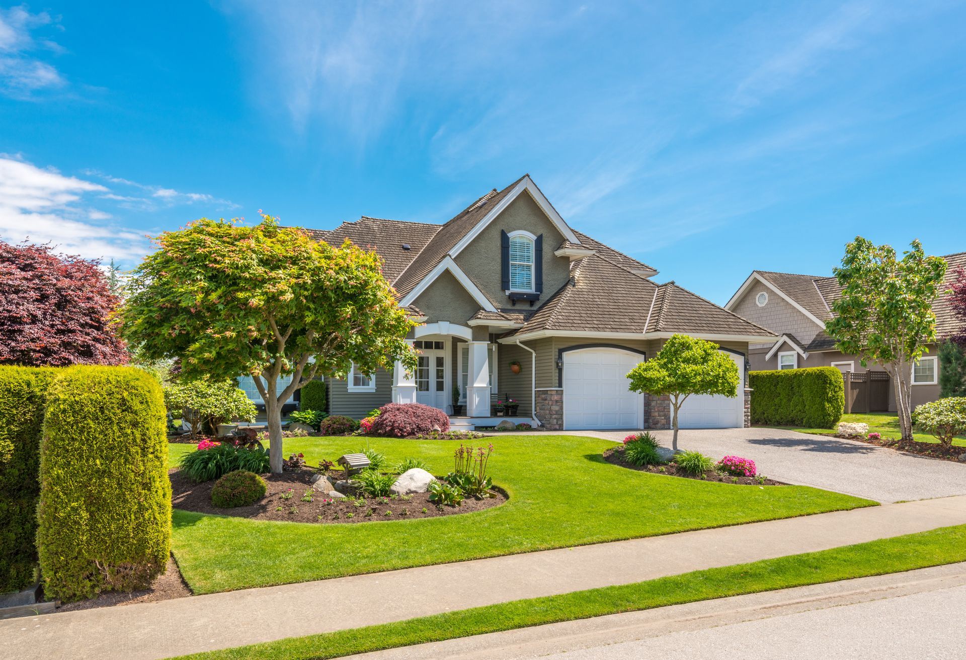 A well-maintained house with a green lawn, trees, and blue sky. The house is gray with a brown roof and has a two-car garage.