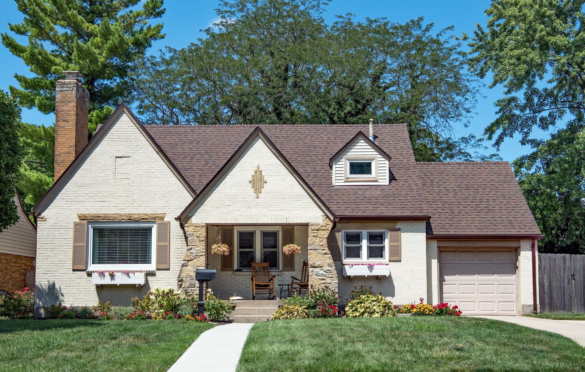 Charming light-colored house with a brown roof, chimney, and garage. Features a small porch, green lawn, and a blue sky.