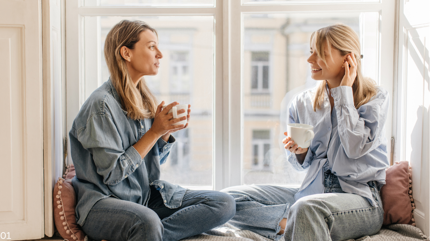 Zwei Frauen unterhalten sich auf einem gepolsterten Fensterplatz und halten Tassen in einem hellen Zimmer mit Blick auf die Stadt.
