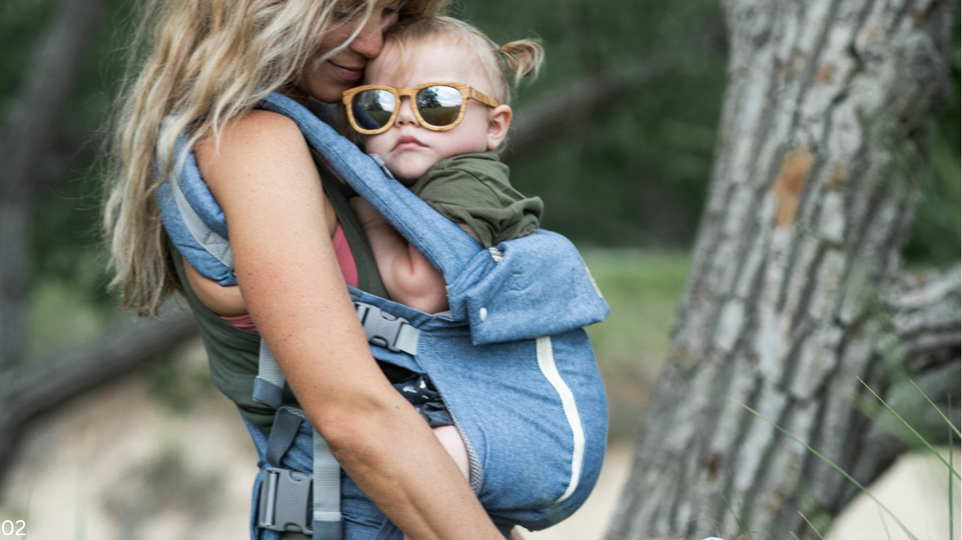 Kind mit Sonnenbrille und grauem Schal, getragen in einer grauen Rückentrage im Freien zwischen Bäumen.
