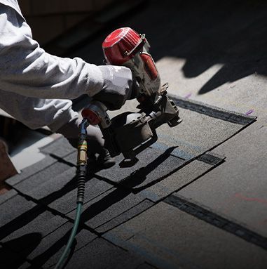 A person uses a nail gun to install roofing shingles on a sunny day.