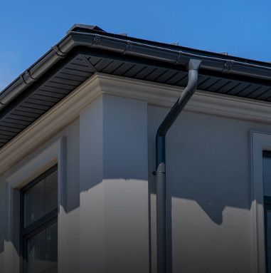 Gray building corner with black gutters, dark trim, and a clear blue sky.