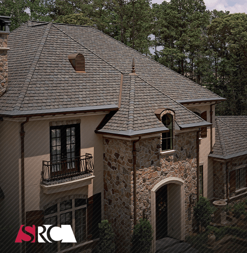 A stone-and-stucco house with a dark shingle roof. It has a turret, dormer and black accents, with trees in the background.