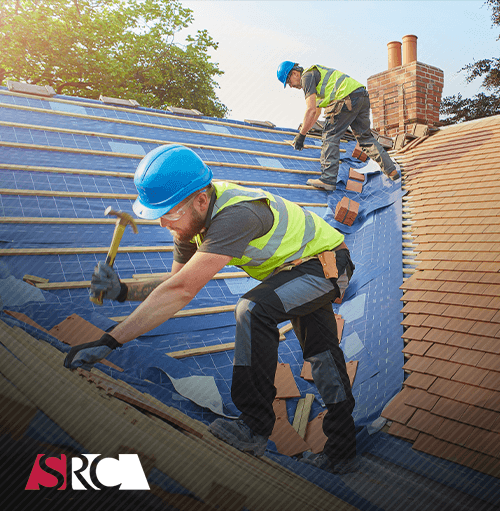 Two roofers in blue helmets and safety vests repair a roof, hammering shingles in sunlight.