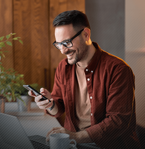 Man in glasses, smiling at his phone indoors, near a laptop and plant.