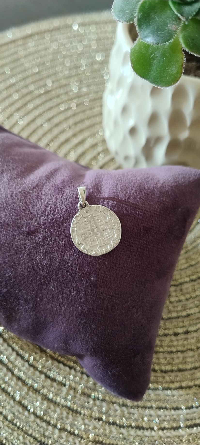 Un colgante circular y brillante sobre un cojín de terciopelo morado, con una planta en una maceta blanca al fondo.