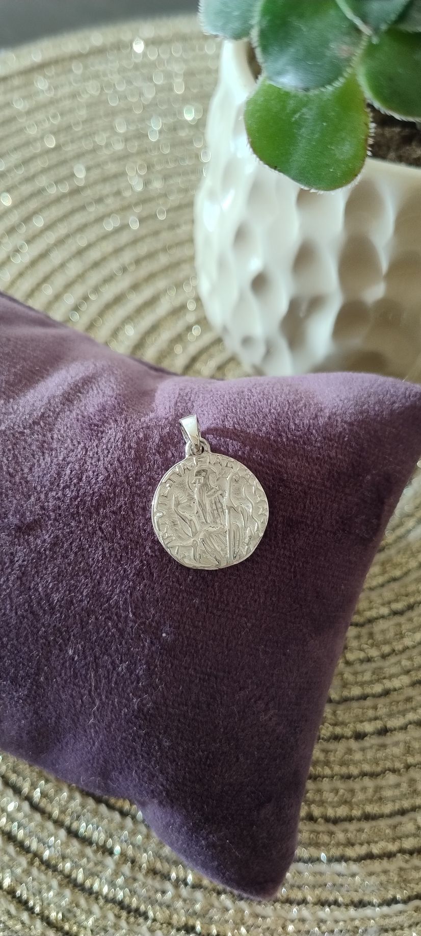 Un colgante circular y brillante sobre un cojín de terciopelo morado, con una planta en una maceta blanca al fondo.