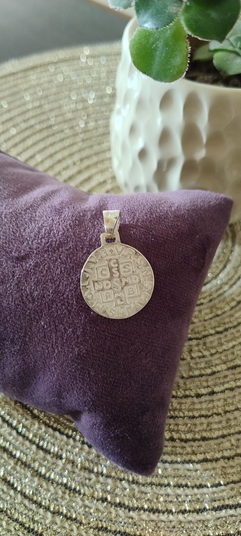 Colgante de plata sobre un cojín morado, con una planta en una maceta blanca al fondo.