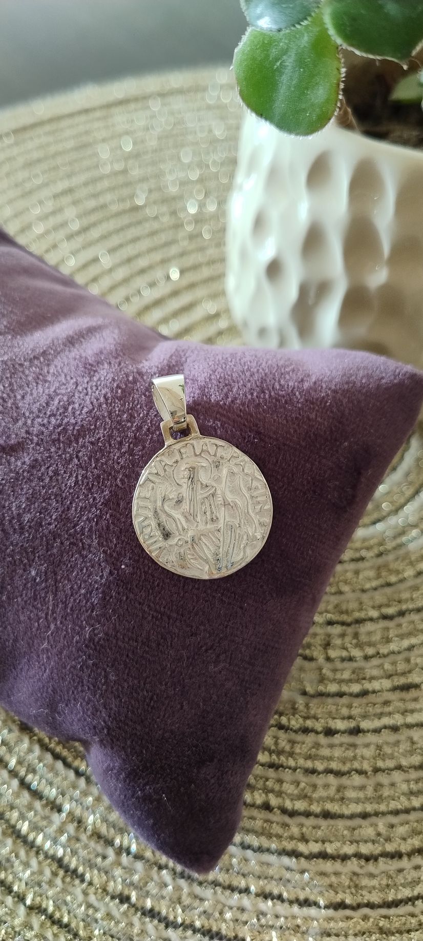Colgante de plata sobre un cojín morado, con una planta en una maceta blanca al fondo.