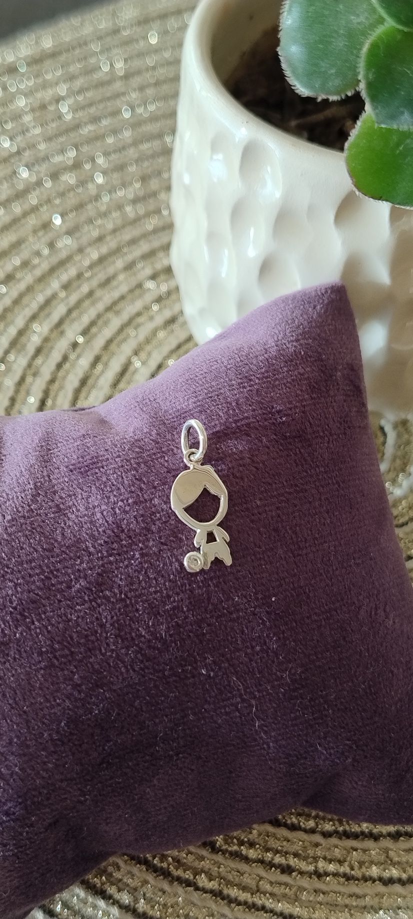 Amuleto de plata con forma de niño sobre una almohada de terciopelo morado, con un jarrón blanco y una planta al fondo.