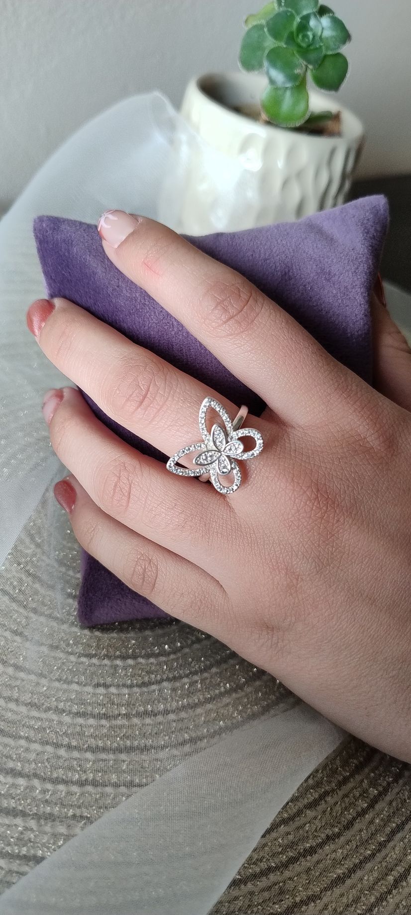 Mano con un anillo de mariposa sobre una almohada morada; pequeña planta al fondo.