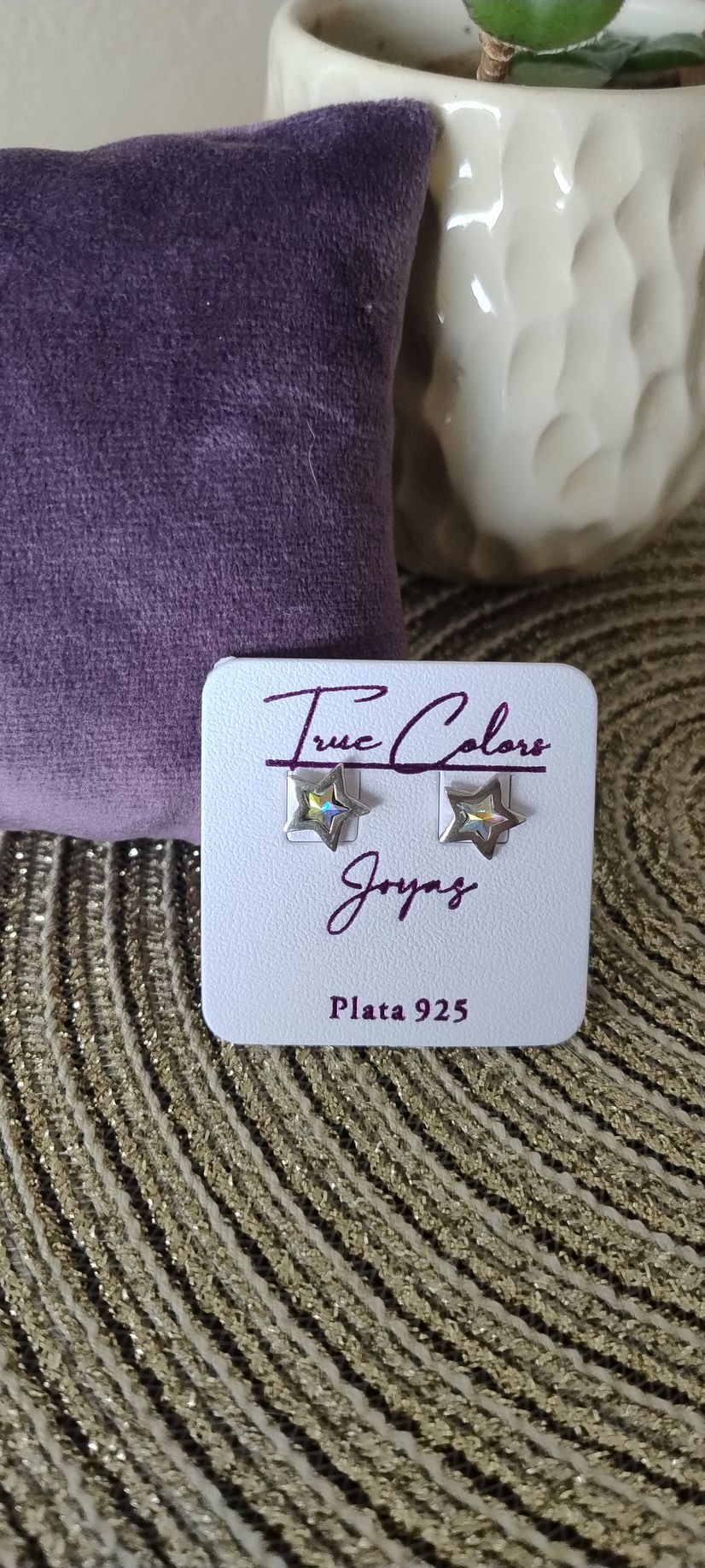 Pendientes de plata en forma de estrella sobre una etiqueta blanca, delante de una almohada morada y una maceta blanca.