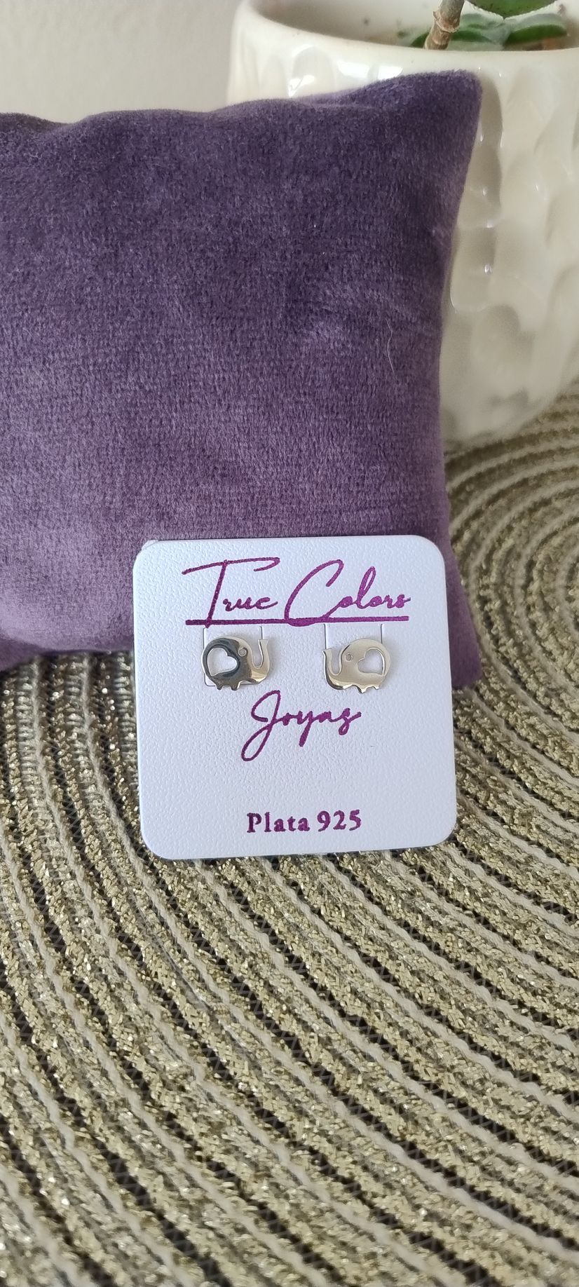 Un par de pendientes expuestos en una tarjeta delante de un cojín morado. La tarjeta dice: 