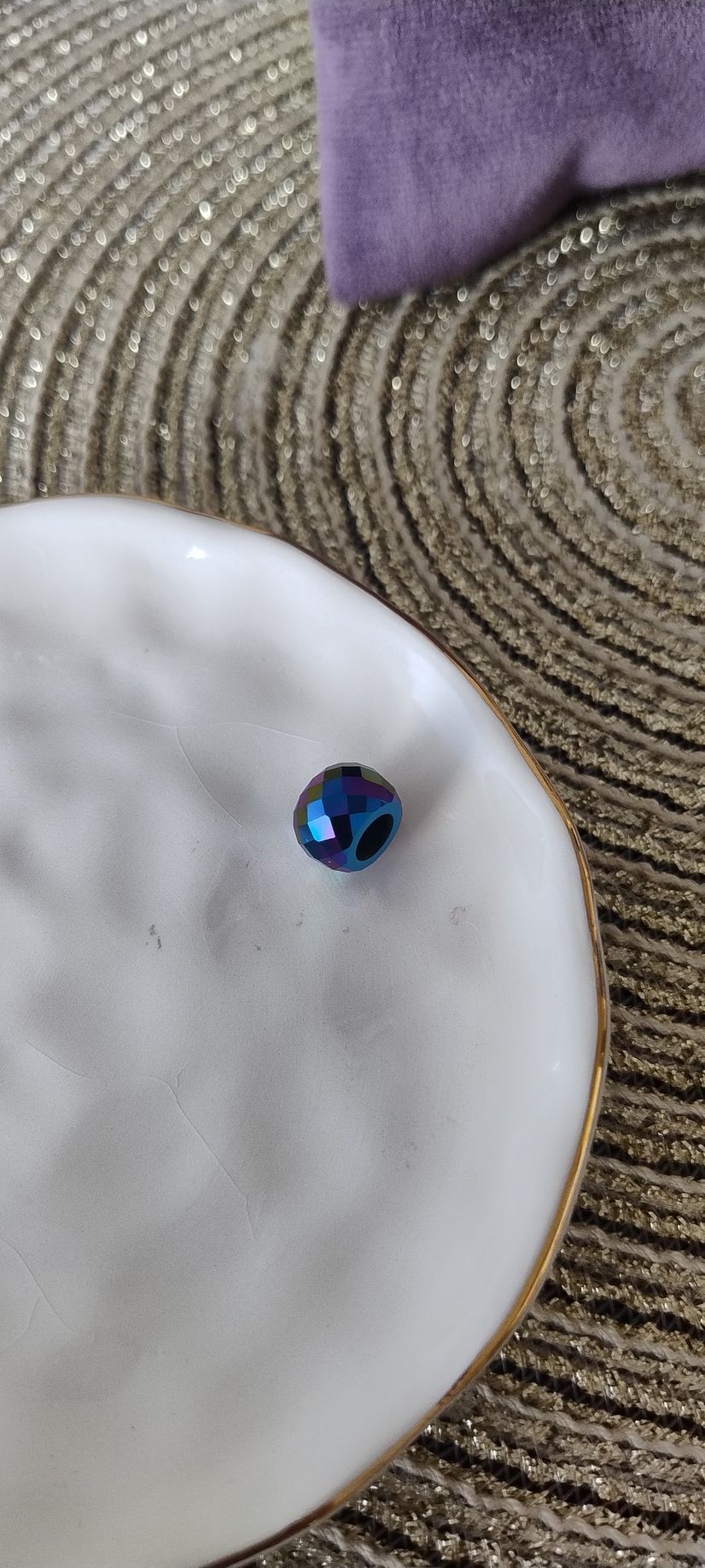Una pequeña cuenta azul reposa sobre un plato blanco texturizado con borde dorado; al fondo se aprecia un cojín morado.